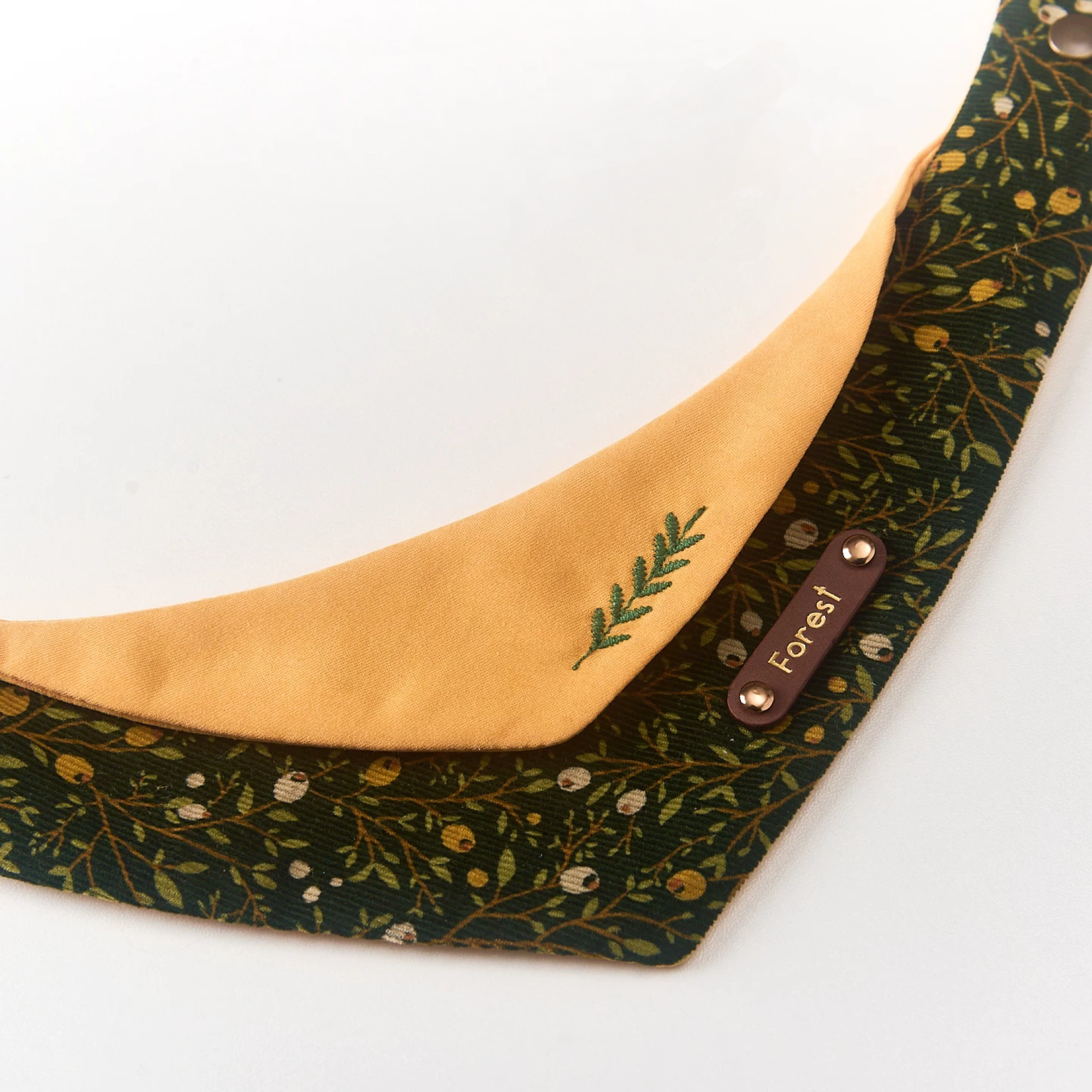 Close-up of embroidered fern sprig on mustard cotton and personalized brown leather name plate against forest botanical corduroy